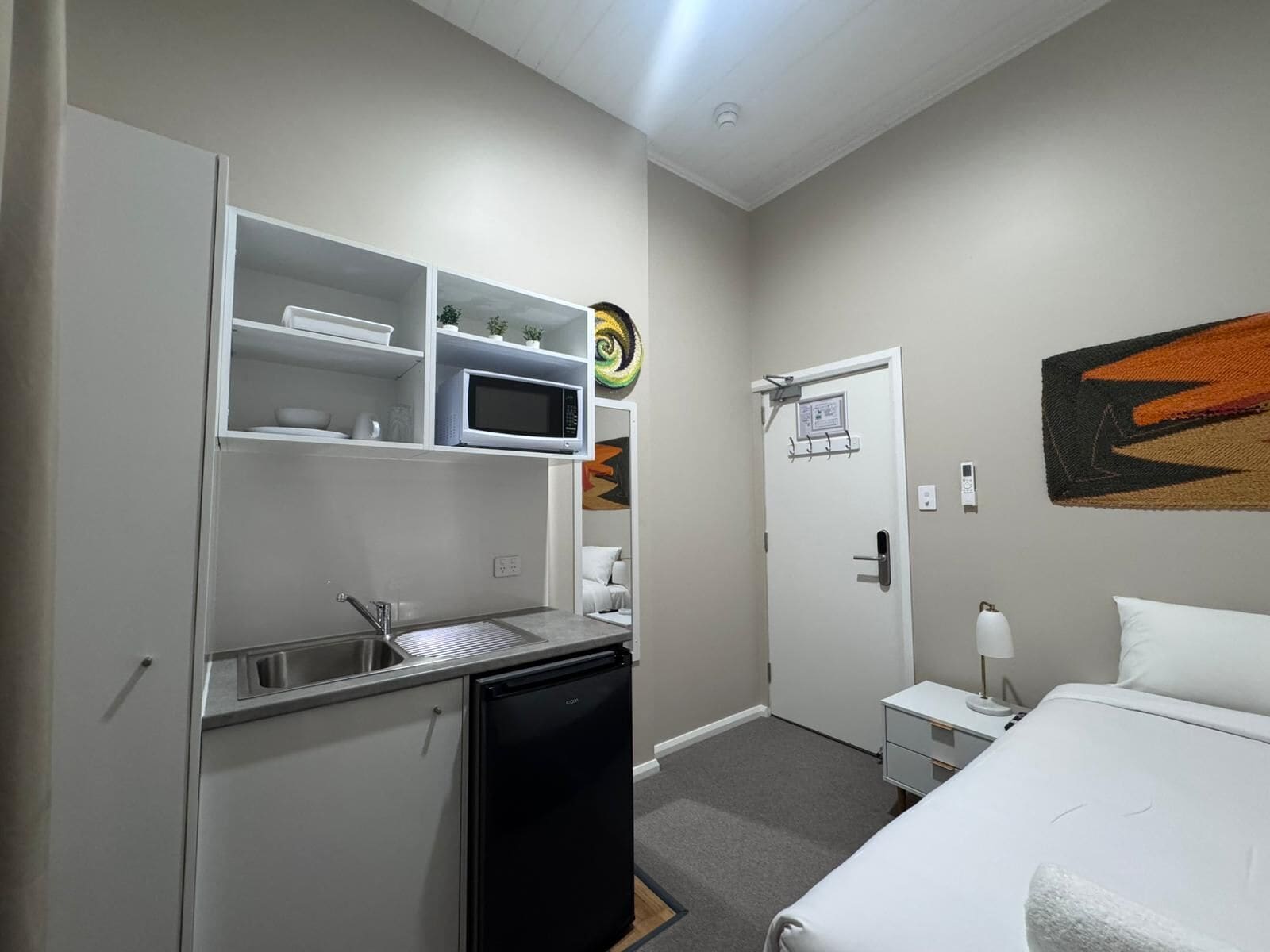 A compact hotel room with a kitchenette, a bed, a bedside table, and a wall-mounted artwork.
