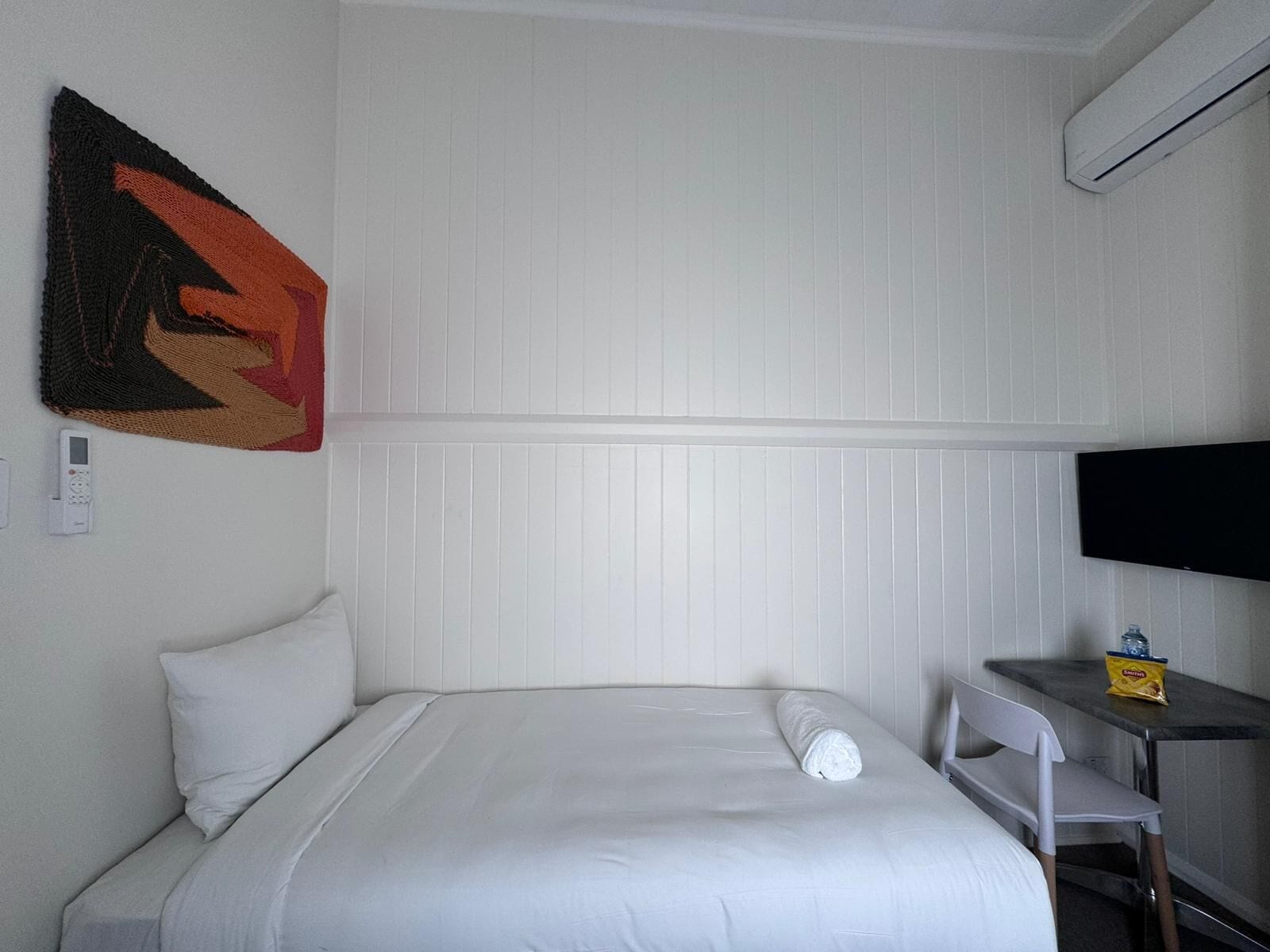 A hotel room with a bed, a wall-mounted TV, a small table with a bag of chips, and a thermostat.