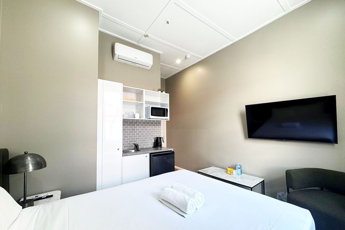 Habitación de hotel con cama, televisión en la pared, una mesita, una silla, microondas, cafetera y kitchenette.