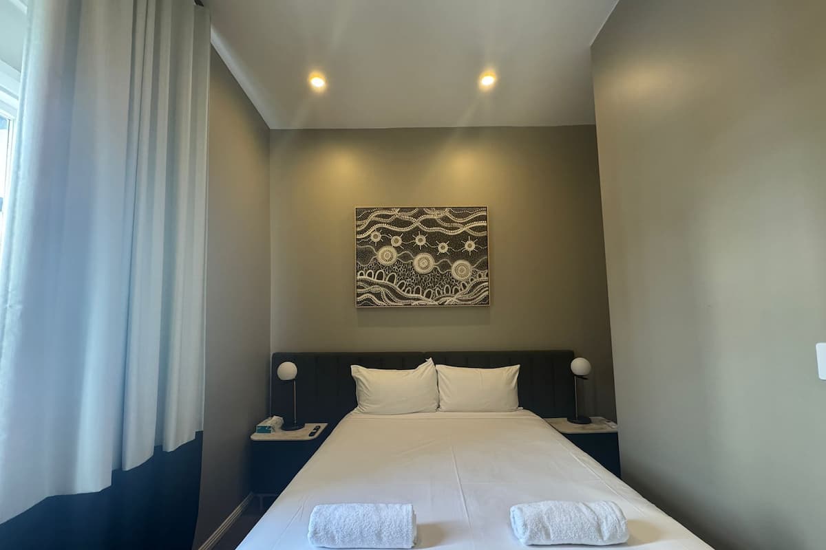 Una habitación de hotel con una cama grande, dos mesitas de noche, una lámpara de noche, una obra de arte en la pared y una ventana con cortinas.