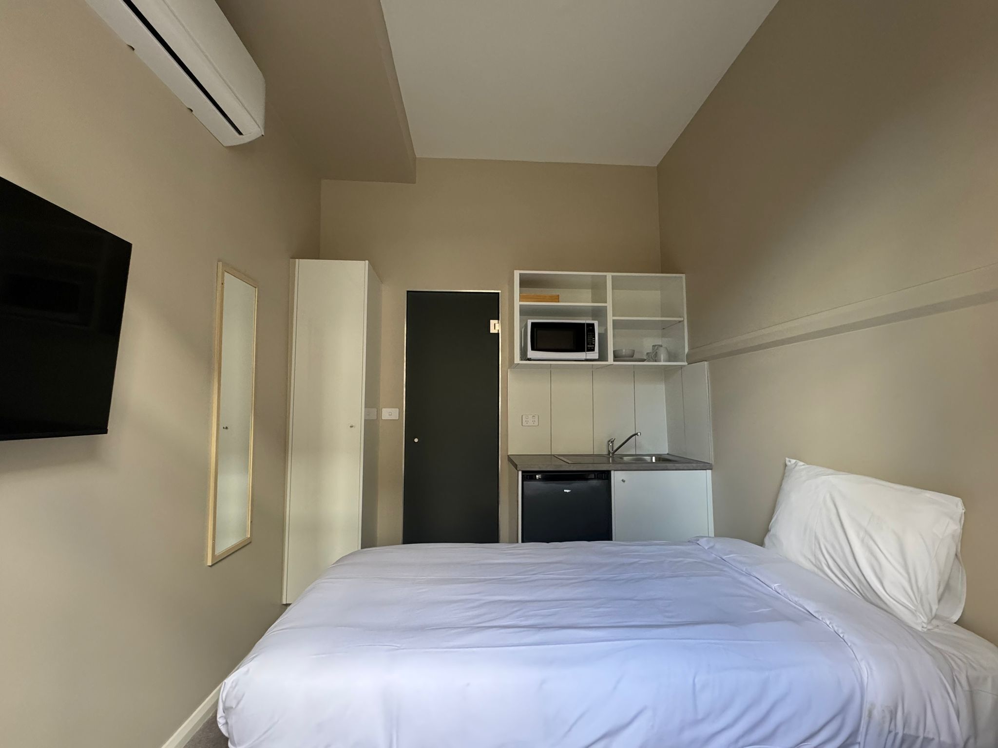 Una camera d'albergo con un letto, una TV a schermo piatto fissata al muro, un angolo cottura con un forno a microonde e un lavandino.