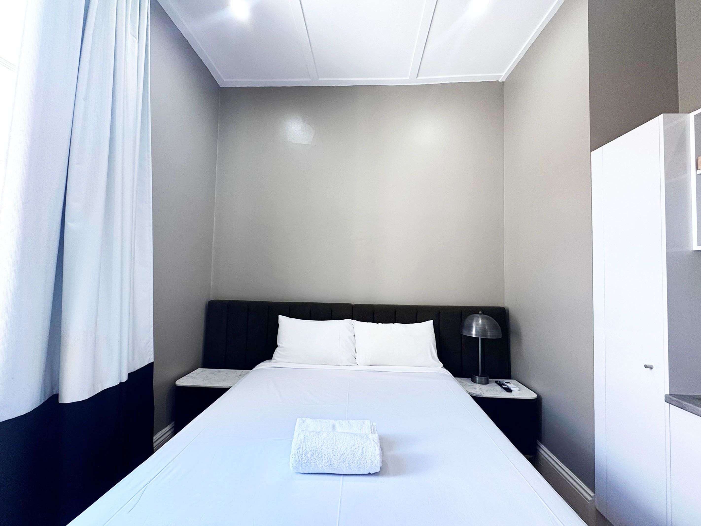 A hotel room with a large bed, white bedding, a black headboard, a nightstand with a lamp, and a wardrobe.