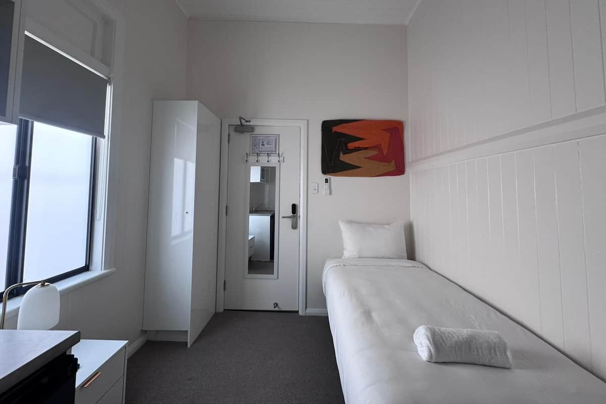 Una habitación pequeña y funcional con una cama individual, un escritorio y un armario. Se ve una puerta que da a un baño con un espejo y una toalla colgada en la pared.
