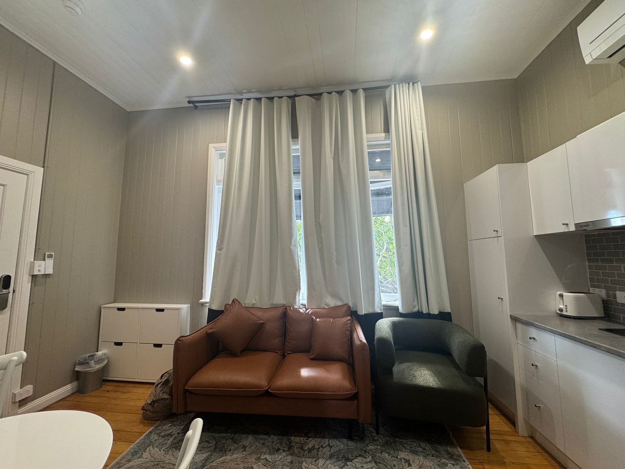 A compact living space with a kitchenette, a brown leather sofa, a dark armchair, and a window with white curtains.