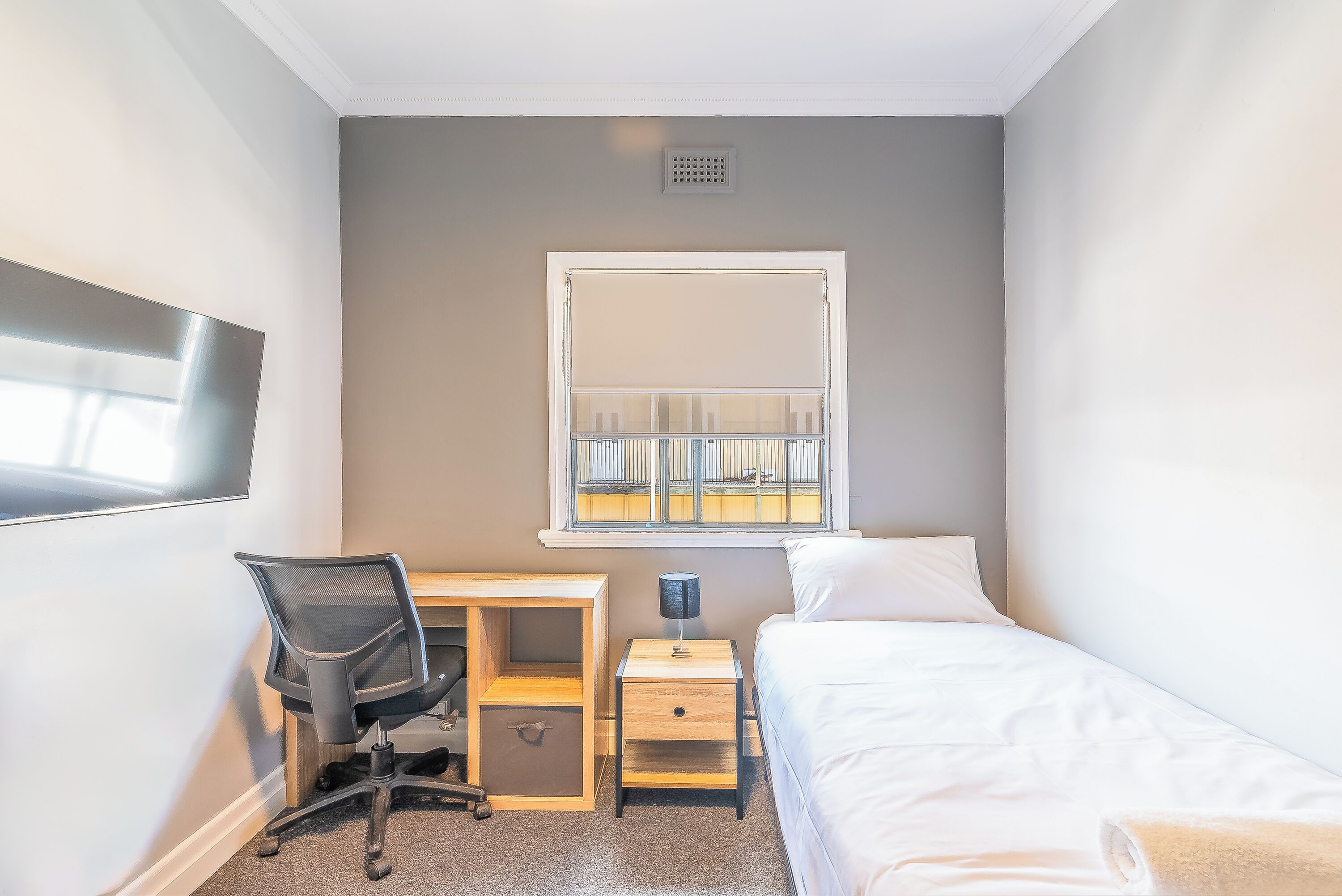 A bedroom with a single bed, a desk with a chair, a small bedside table with a lamp, and a window with blinds.