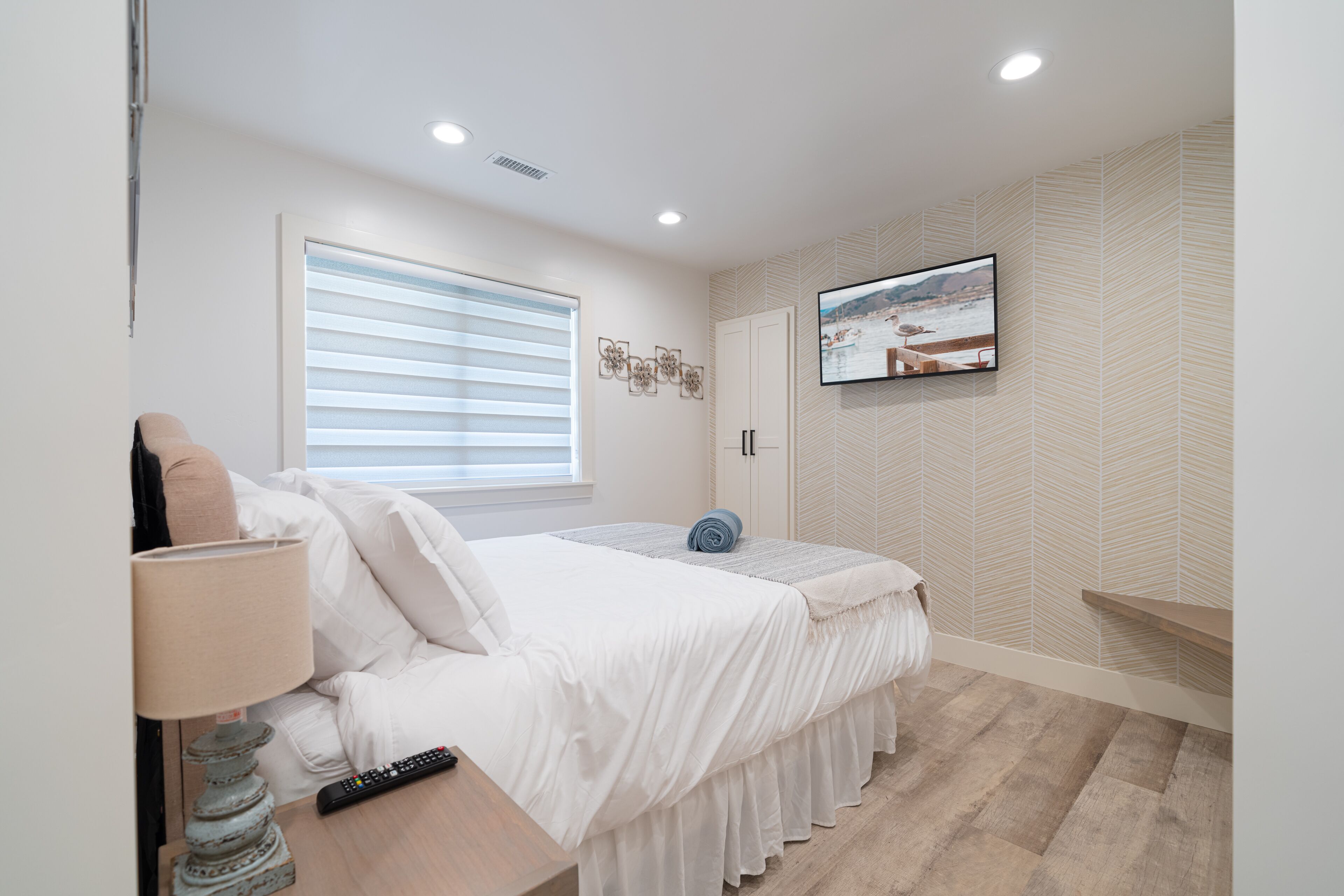 A bedroom with a bed, a TV mounted on the wall, a window with blinds, and a wooden floor.