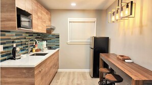 Cocina compacta con armarios de madera, un lavamanos de acero inoxidable y un refrigerador negro. Hay una mesa de comedor de madera con dos taburetes y una lámpara colgante encima.