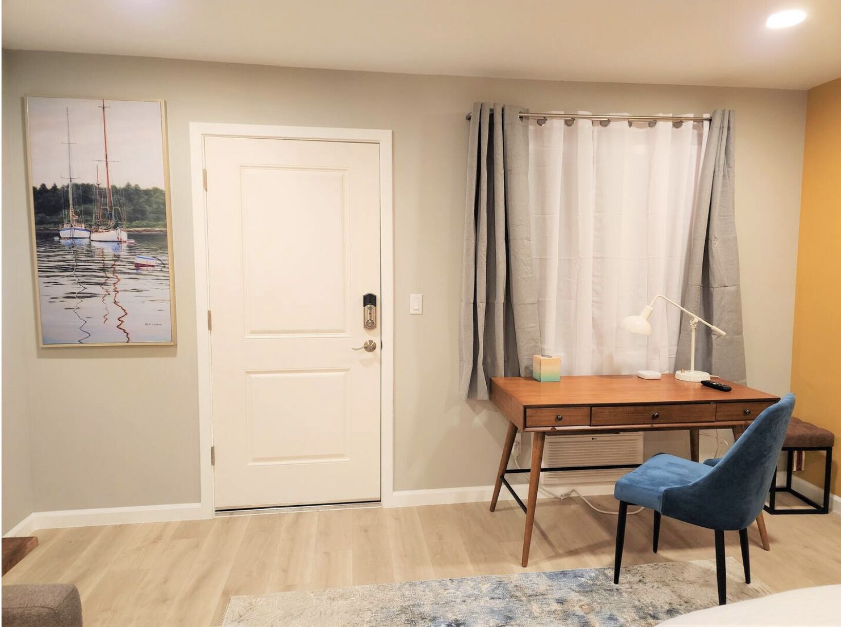 Una habitación con un escritorio de madera, una silla, un cuadro, una puerta y una ventana con cortinas.