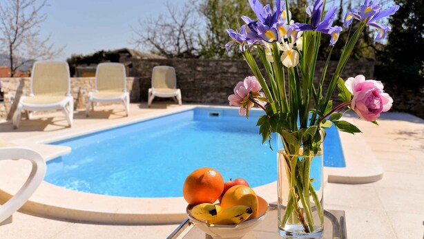 Escena junto a una piscina con aguas cristalinas, sillas de descanso, un florero con flores y frutas.