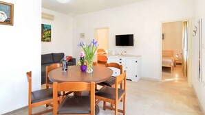 Un comedor con una mesa redonda de madera, cuatro sillas, un florero con flores, un sofá, un televisor y una cama al fondo.