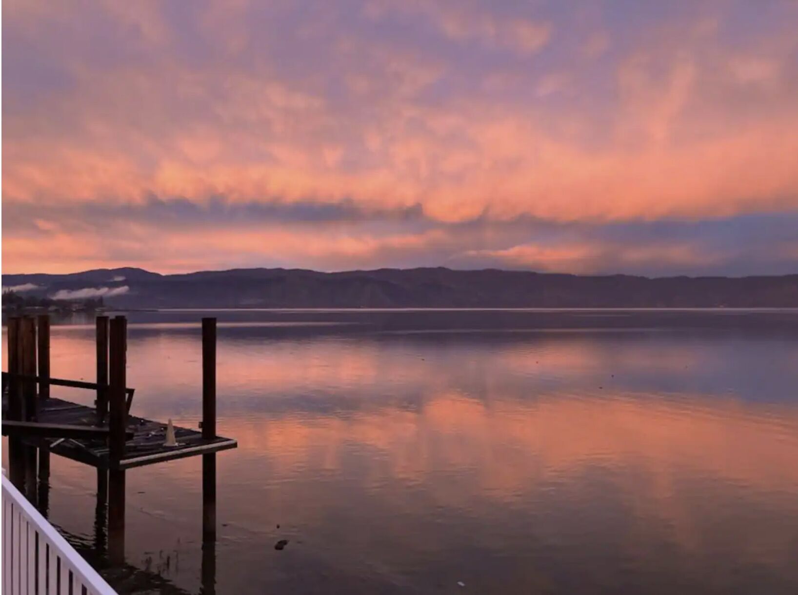 A calm lake with a wooden dock and a distant mountain range under a colorful sky.