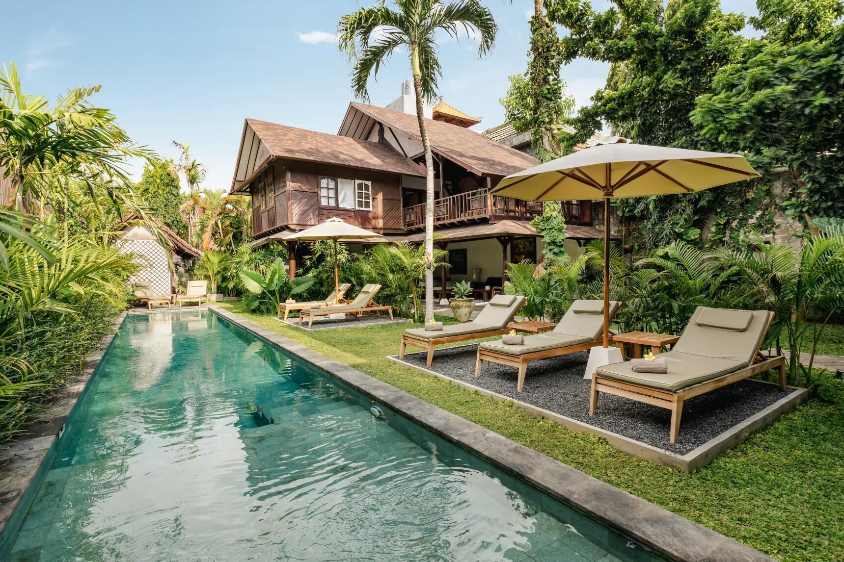 Un espace aménagé au bord de la piscine, avec des chaises longues et des parasols, entouré d’une végétation luxuriante et d’une grande maison dotée d’un balcon.