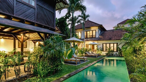 A poolside area with lounge chairs and umbrellas, surrounded by lush greenery and a swimming pool. - Legian 6BR Villa 16px Big Group Walk to 66 Beach (Bali)