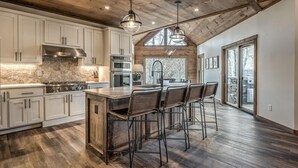 A spacious kitchen with white cabinets, a large island with bar stools, a stone backsplash, and a high ceiling with exposed beams. - Mountain Brooks Retreat - Hot Tub | Close to Downtown Blue Ridge | Outdoor Fireplace (Blue Ridge)
