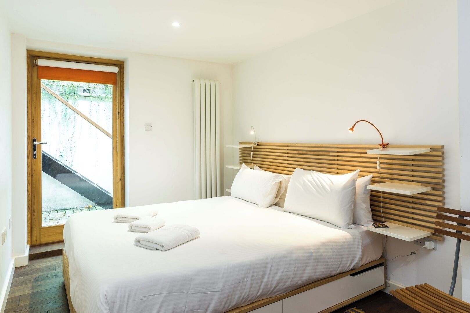 Een slaapkamer met een bed, kussens, een nachtkastje, een lamp en een raam met uitzicht op een trap.