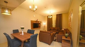 A modern living room with a wooden dining table, blue chairs, a brown sofa set, and a TV mounted on the wall. - Two Bedroom Apartment Near NMC Sociality Hospital (Dubai)