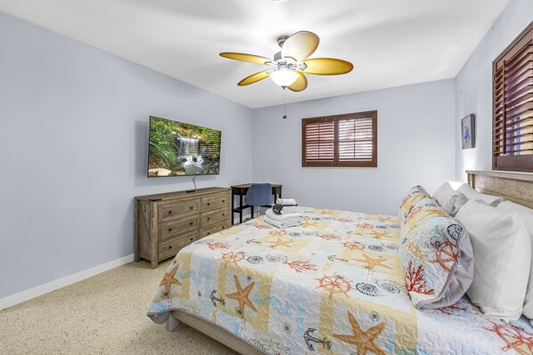 Un dormitorio con cama, ventilador de techo, televisor, cómoda de madera, una silla y ventana con persianas.