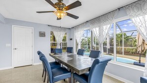A dining room with a large table, blue chairs, a ceiling fan, and large windows with curtains.