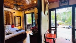 A hotel room with a bed, a desk, a chair, a ceiling fan, and a view of the street outside. - Anisa Mekong Riverside Villa (Luang Prabang)