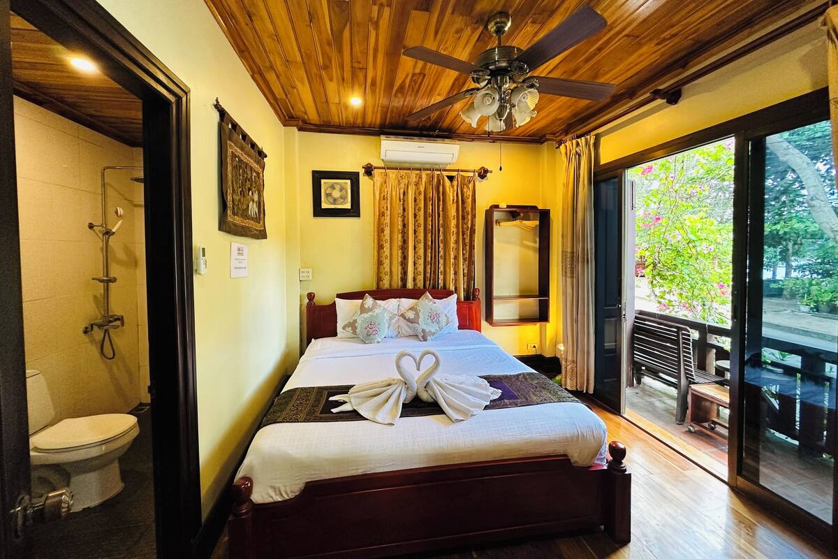 A hotel room with a wooden ceiling, a bed with white bedding and a towel, a shower, a large mirror, and a balcony with a view of trees.