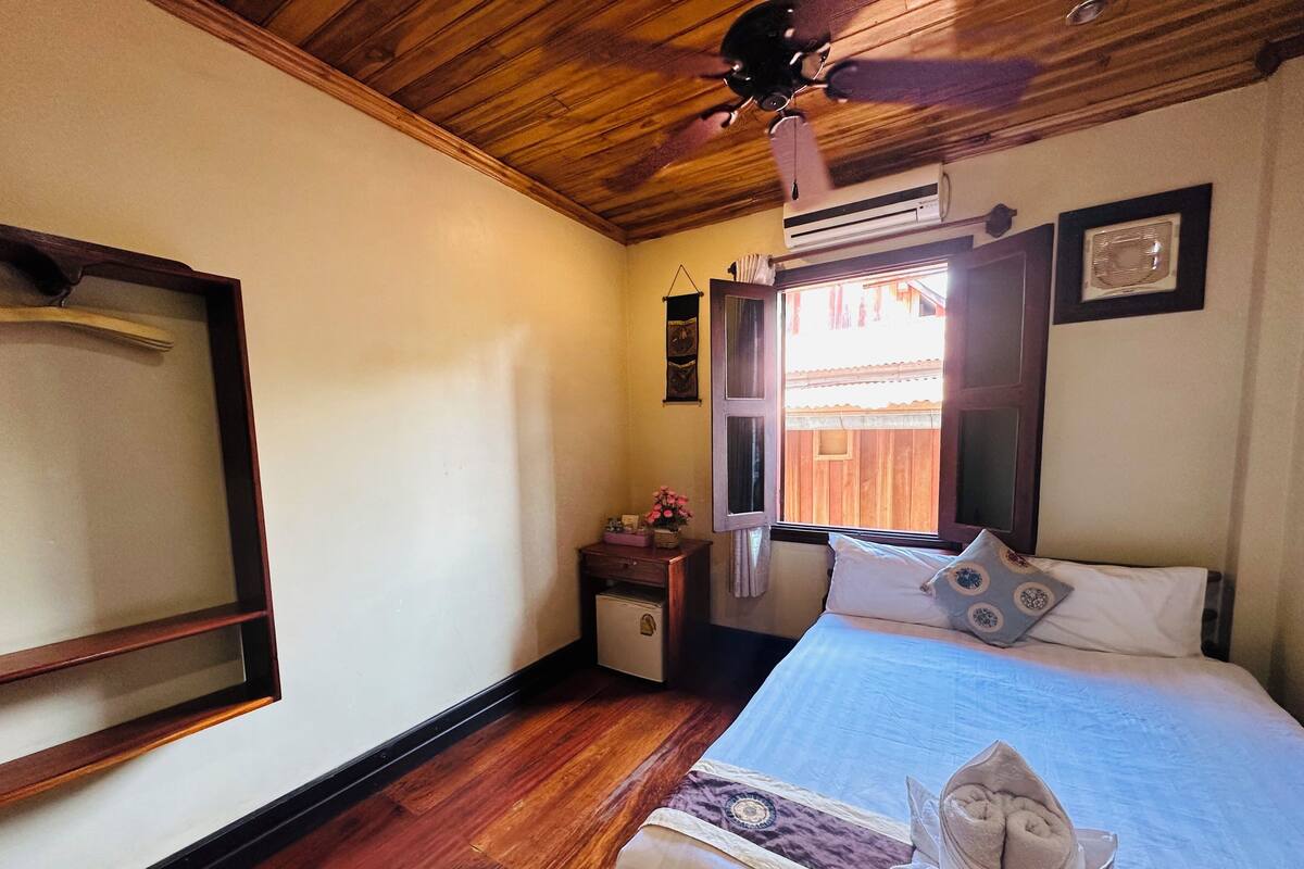 A bedroom with a wooden ceiling, a bed with a blue bedspread, a window with curtains, a small bedside table with a vase of flowers, and a ceiling fan.
