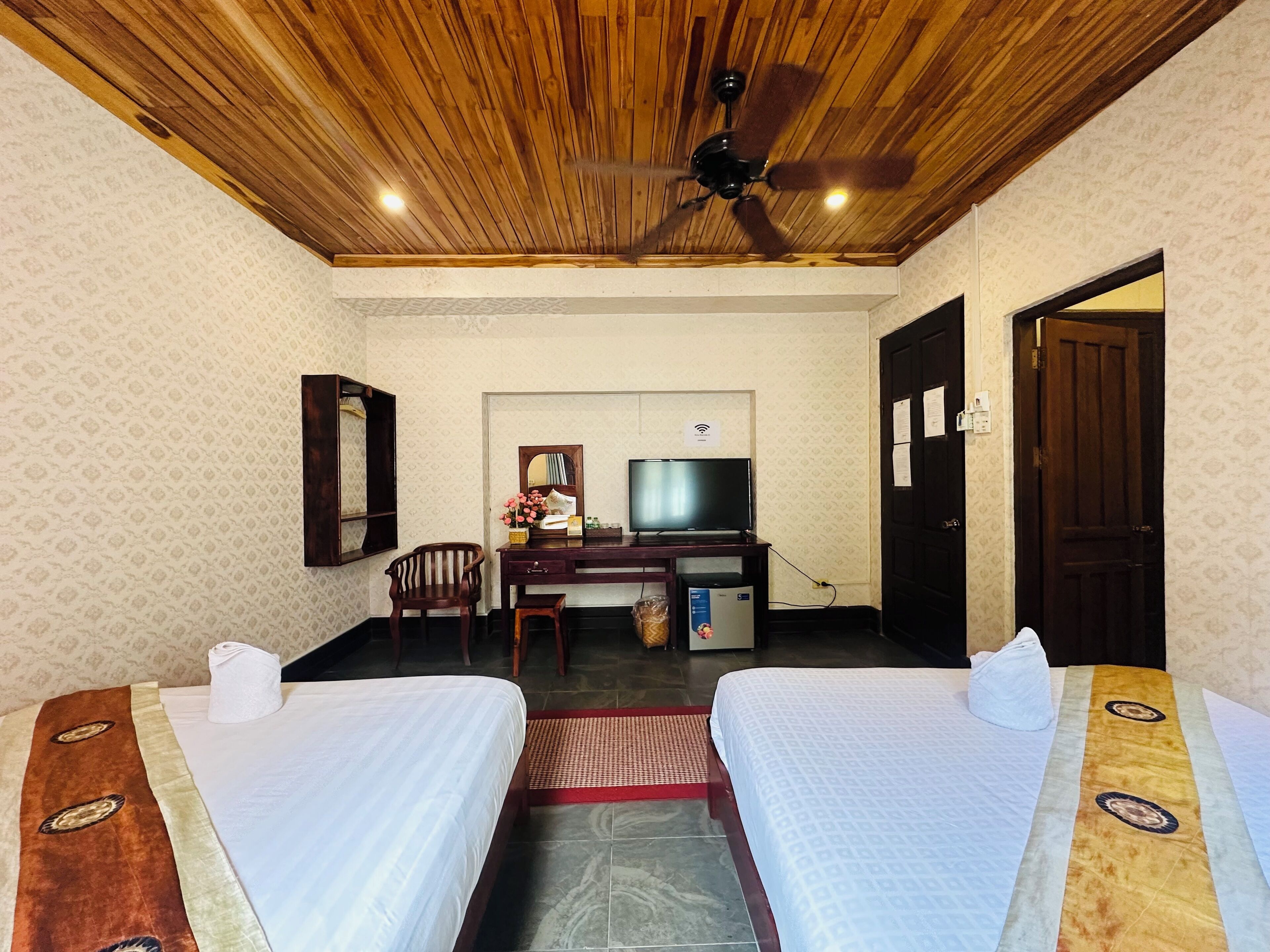 Habitación de hotel con dos camas, un escritorio con televisor, ventilador de techo y techo de madera.