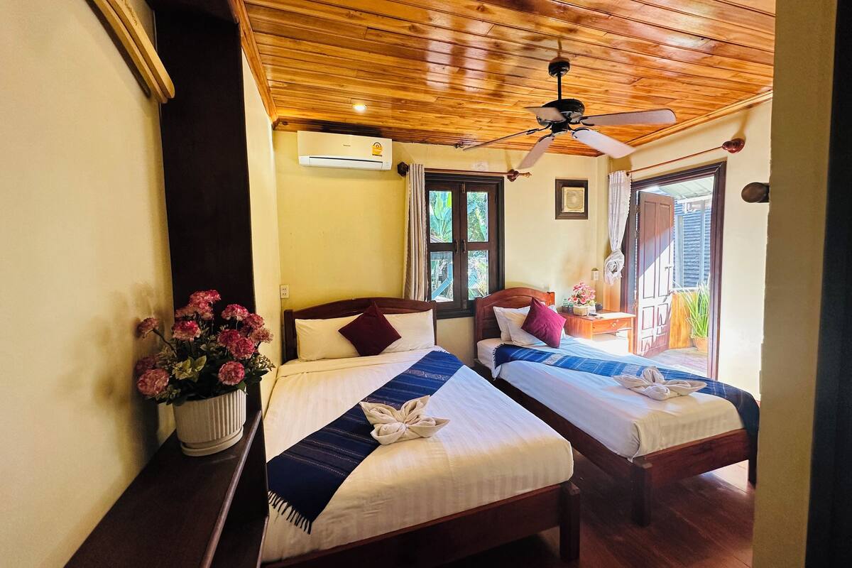 A hotel room with two beds, wooden ceiling, ceiling fan, air conditioning unit, window with curtains, and a small table with a plant.
