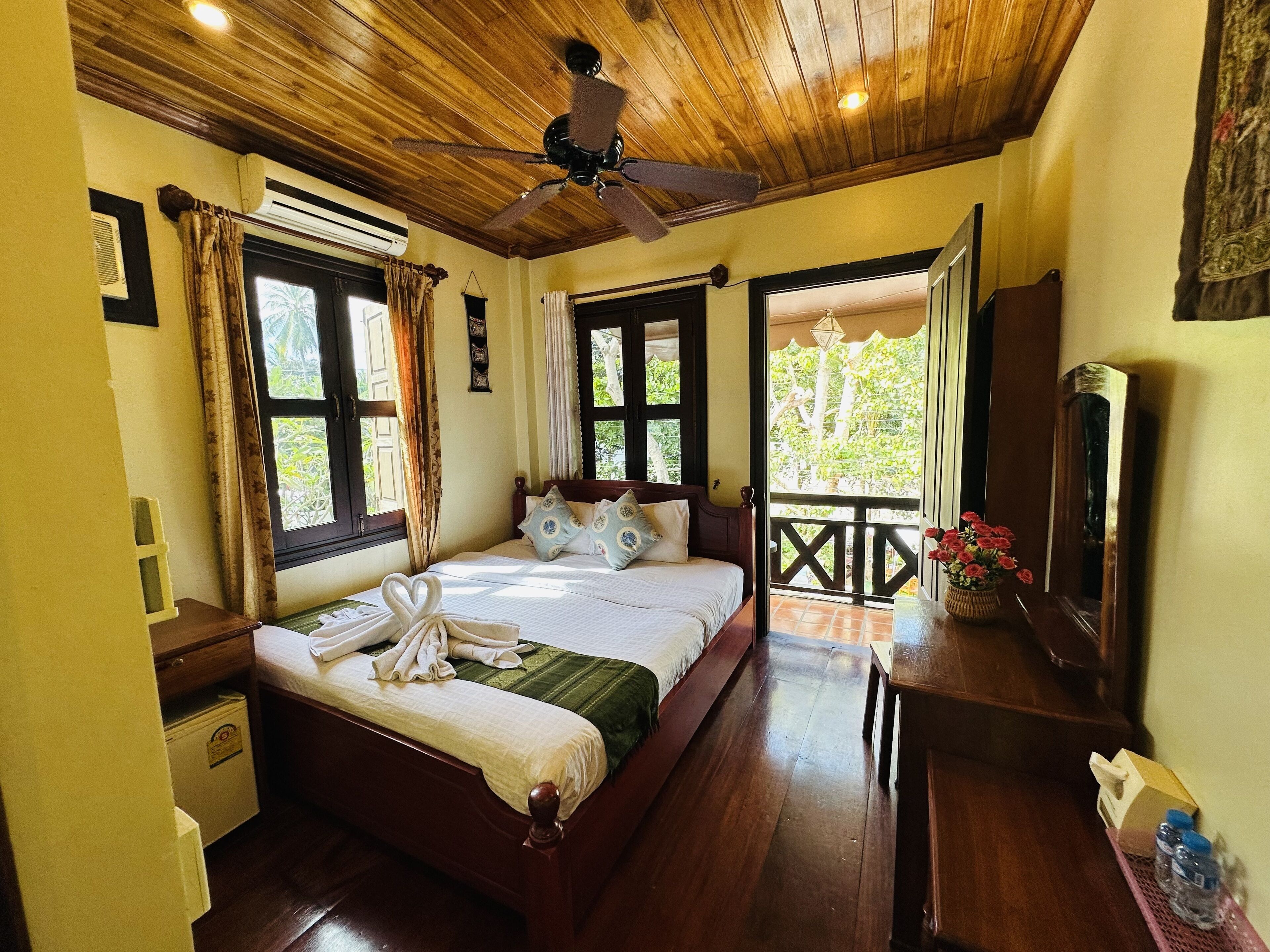 Un dormitorio con techo de madera, una cama con manta verde, una mesita de noche de madera, un aparador, un espejo y una ventana con cortinas.