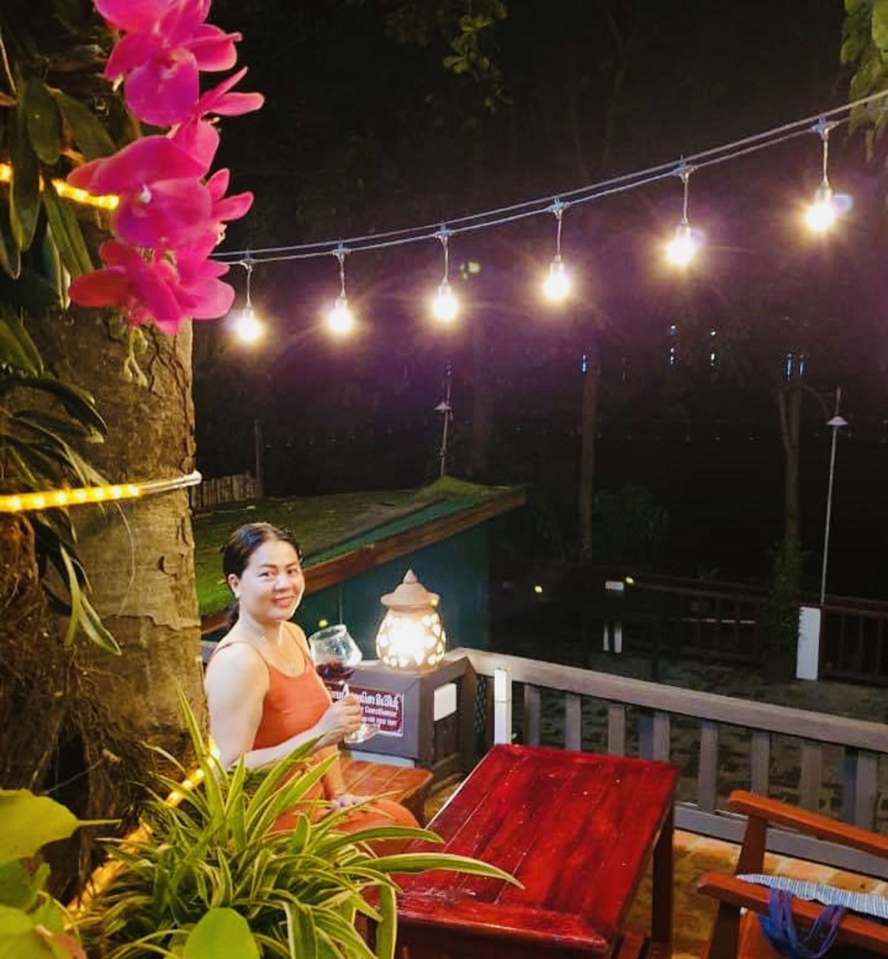 A person seated at a table with a drink, under string lights, with a view of a building and trees in the background.