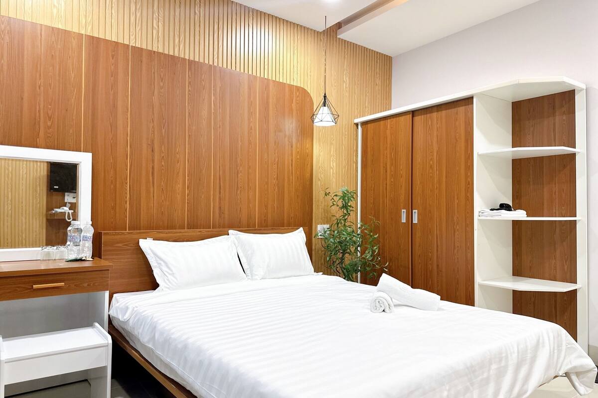 A modern bedroom with a wooden panel wall, a bed with white bedding, a bedside table with a mirror, a hanging light fixture, a potted plant, and a wardrobe with shelves.