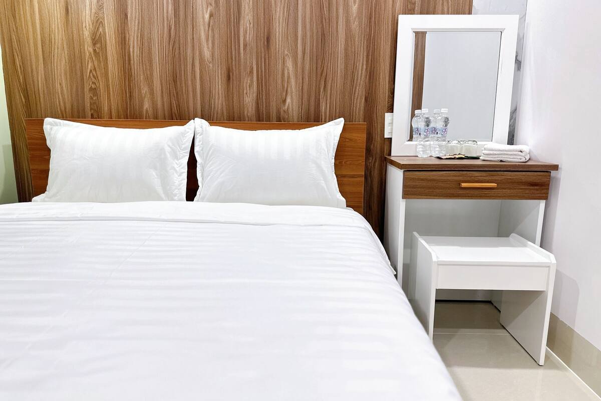 A neatly made bed with white linens, a wooden headboard, a small wooden nightstand with a mirror, and a white bench.
