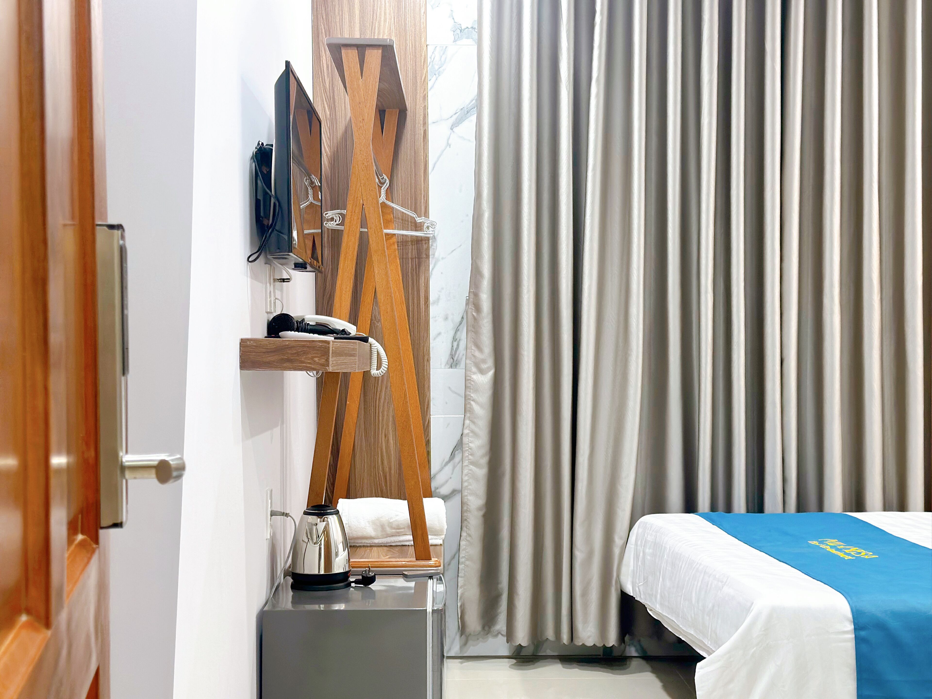 A hotel room with a bed, a desk with a telephone and a kettle, a wooden TV stand, and a window with curtains.