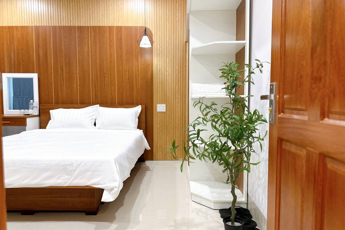 A hotel room with a wooden headboard, a bed with white bedding, a potted plant, and a white wardrobe with shelves.