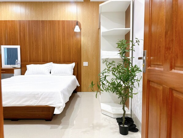 A hotel room with a wooden headboard, a bed with white bedding, a potted plant, and a white wardrobe with shelves. - Milanesa Hotel & Apartment (Ho Chi Minh City)