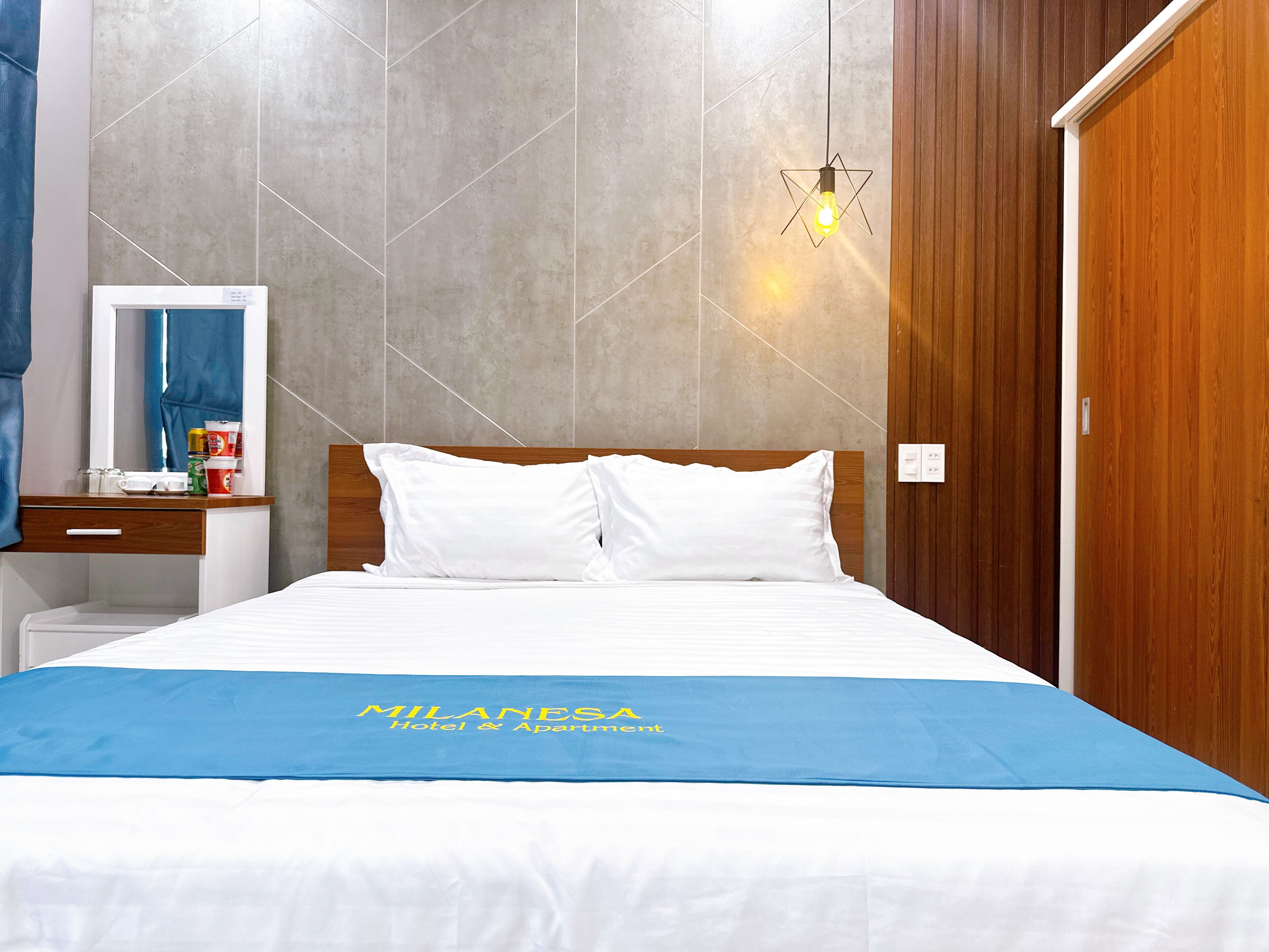 Une chambre d’hôtel avec un lit, une tête de lit en bois, une table de chevet avec un miroir et un luminaire fixé au mur.
