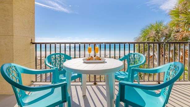 A balcony with turquoise chairs and a table set for two, overlooking a beach.