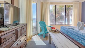 A hotel room with a wooden dresser, a blue chair, a bed with a blue quilt, and a window with curtains.