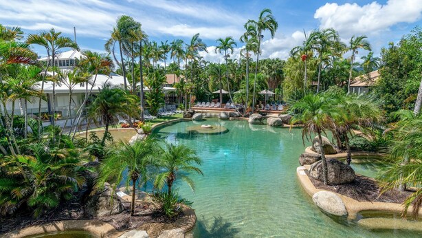 A resort with a clear pool surrounded by palm trees and lounge chairs.