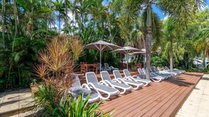 A wooden deck with sun loungers and umbrellas, surrounded by palm trees and tropical plants.
