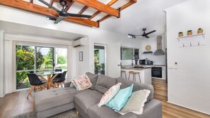 A modern living room with a grey sectional sofa, a dining area with a glass table, and a kitchen with white cabinets and a black stove.