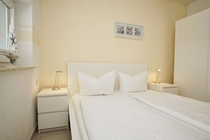A neatly arranged bedroom with a bed, two bedside tables, a nightstand, a window with curtains, and a framed picture on the wall.