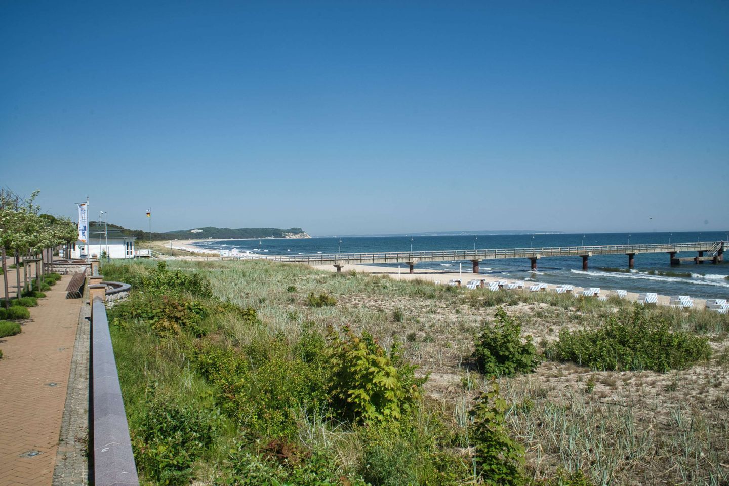 A coastal walkway with a bridge over the water, a beach, and a building with a flag.