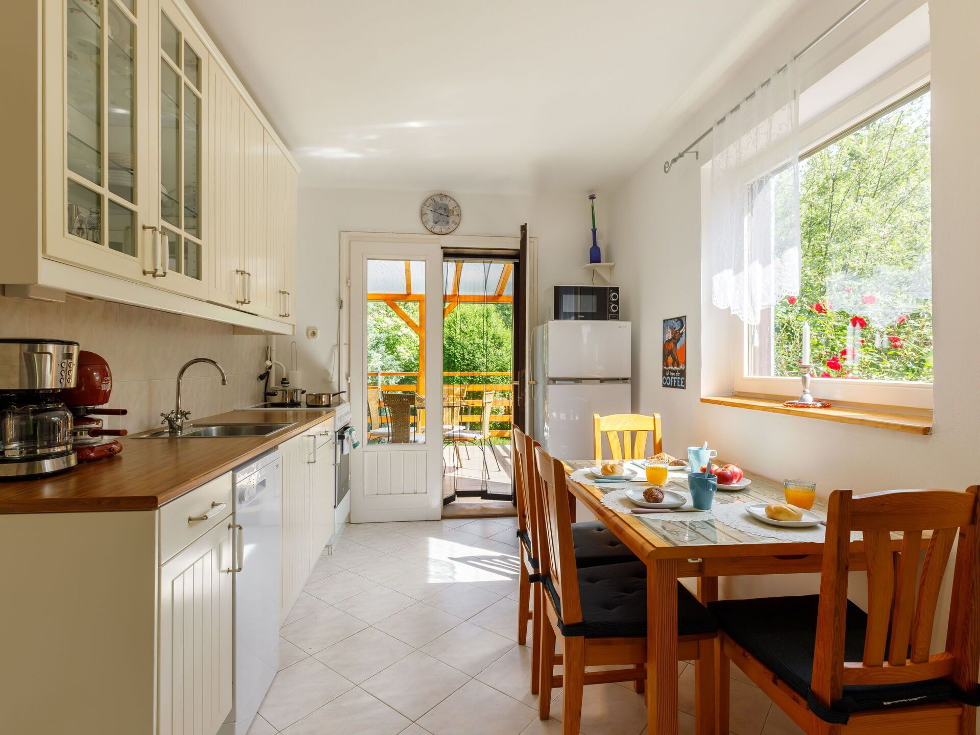 Une cuisine avec des meubles blancs, une table à manger avec des chaises, un réfrigérateur et une fenêtre donnant sur un espace verdoyant.
