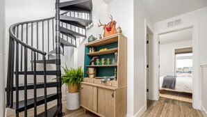A modern interior with a spiral staircase, a wooden cabinet with decorative items, and a view into a bedroom with a bed and a window. - The Loft on Gillis | AC | Downtown | Beach | Rooftop | (Traverse City)