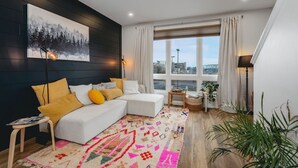 A modern bedroom with a white sofa, colorful rug, and a large window with curtains. - The Loft on Gillis | AC | Downtown | Beach | Rooftop | (Traverse City)
