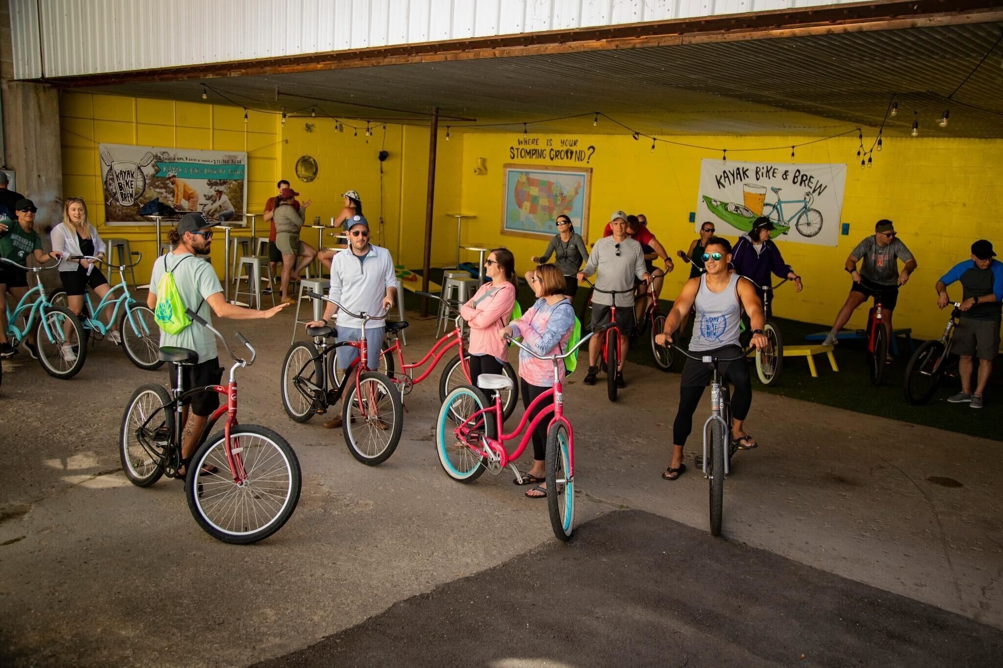 A group of people on bicycles in an indoor area with yellow walls, some holding drinks, and a banner that reads "Kayak Bike & Brew".