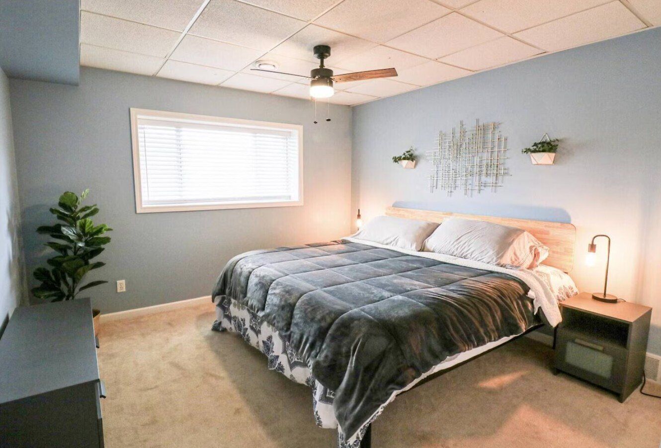 A bedroom with a bed, a ceiling fan, a nightstand, a wall-mounted plant, and a window with blinds.