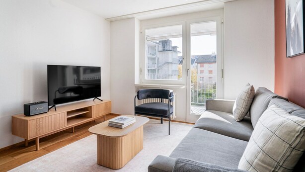 Een moderne woonkamer met een flatscreen televisie op een houten standaard, een zwarte stoel, een grijze bank en een kleine houten salontafel.