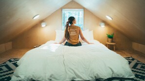 Une personne est assise sur un lit dans une pièce dont le plafond est en pente, avec une fenêtre et une petite plante posée sur une table d’appoint.