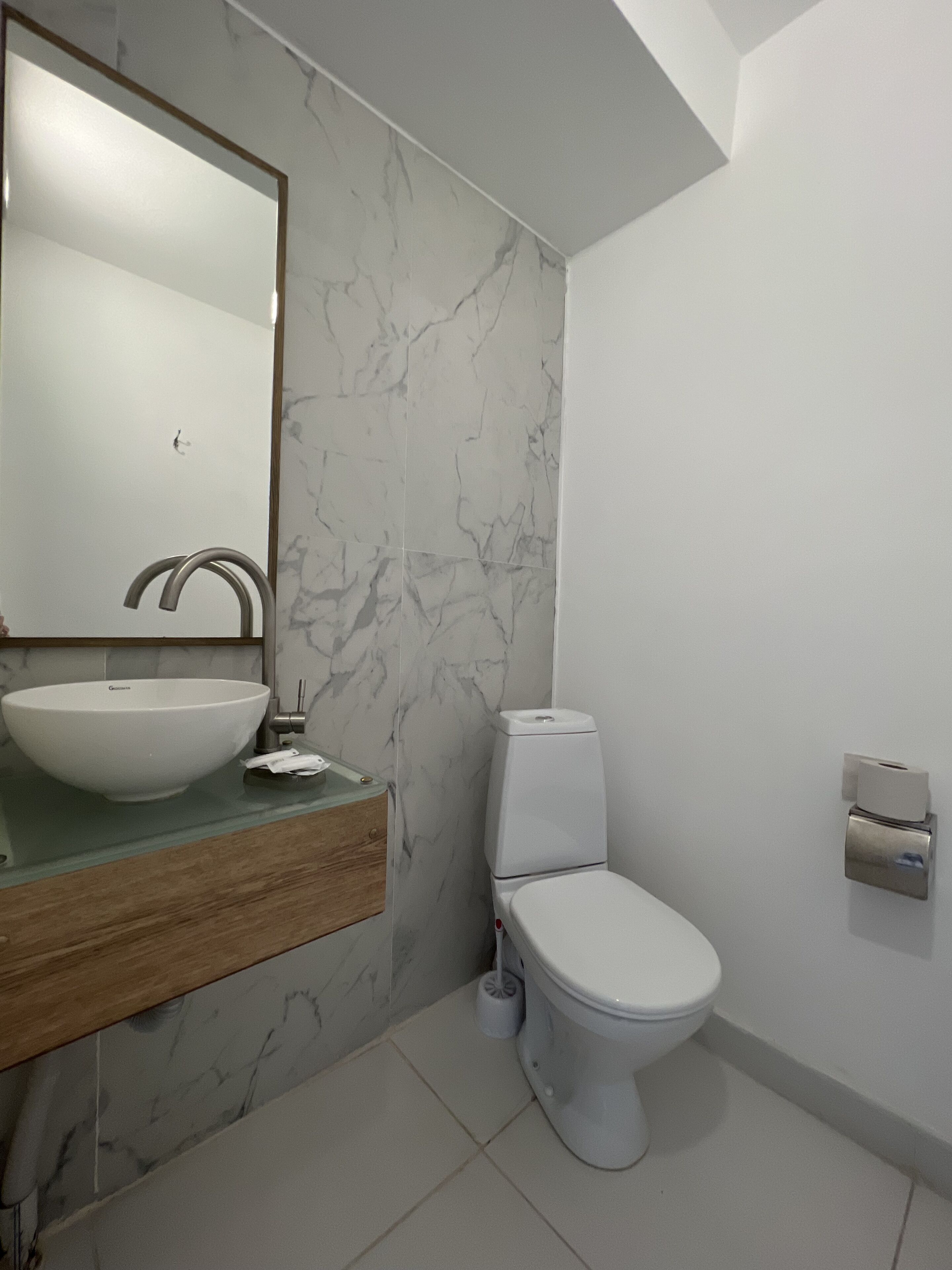 Ein Badezimmer mit Marmorfliesenspiegel, einem Holzbadezimmerschrank, einem weißen Waschbecken, einer Toilette und einem an der Wand montierten Toilettenpapierhalter.